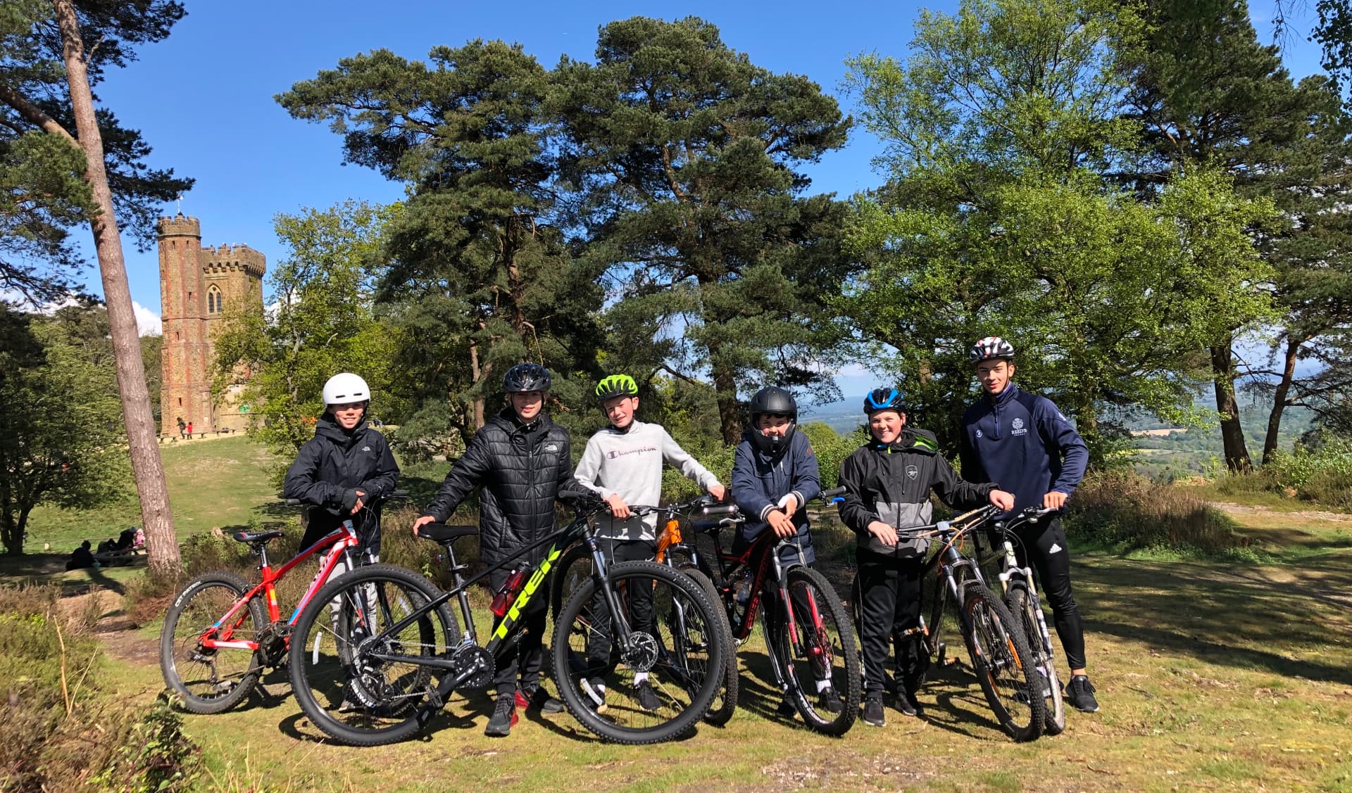 A group of young mountain bikers standing with their bikes on a Surrey Hills viewpoint beside Leith Hill Tower under a blue sky