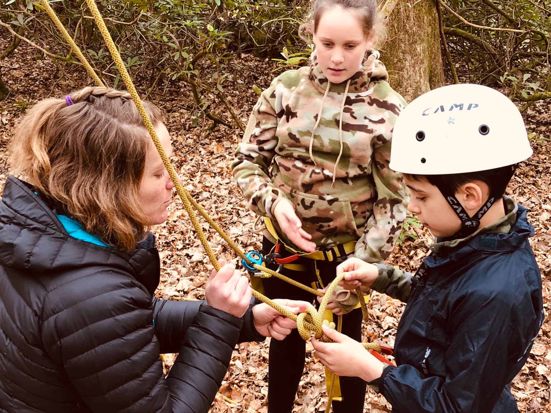 An instructor teaching two young people to tie a knot using climbing rope and a harness in woodland