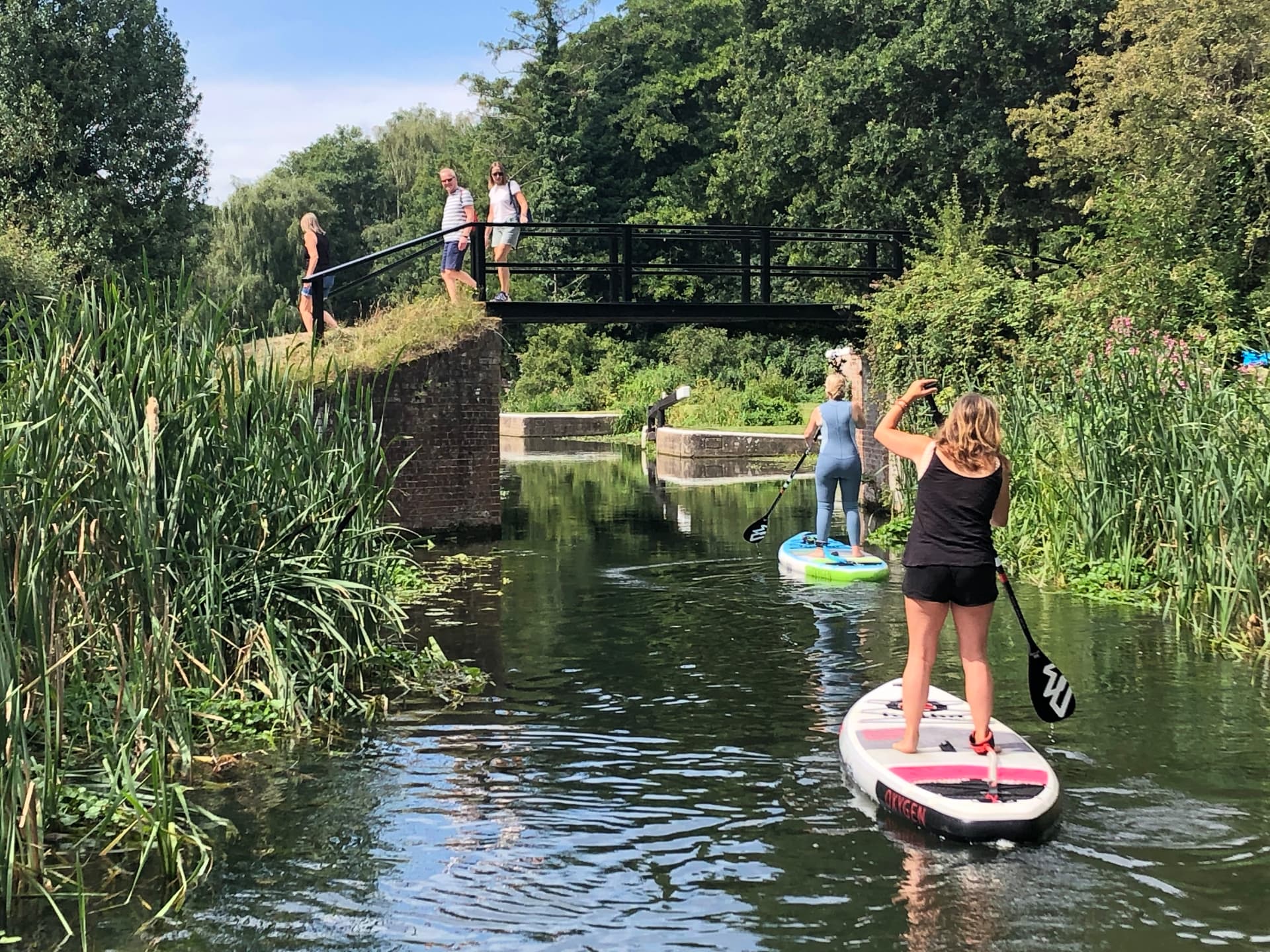 Paddleboarders approaching a lock on the River Wey Navigation as walkers cross the footbridge above