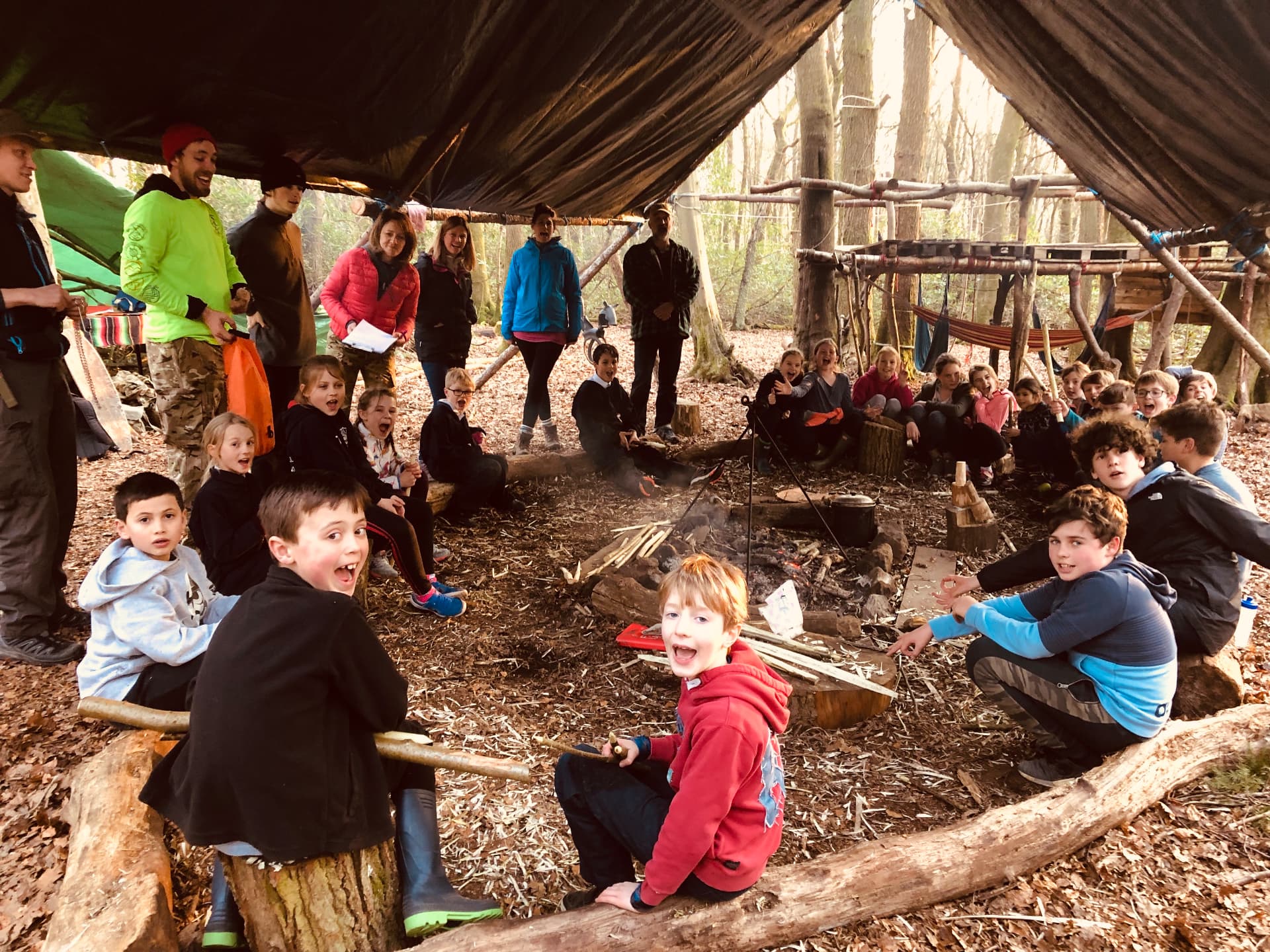 A large group of children and adults gathered around a woodland fire circle beneath a tarpaulin shelter at base camp