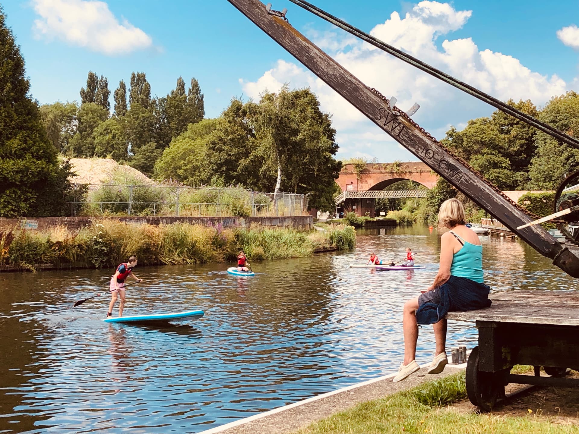 Young paddleboarders on the River Wey Navigation beside a historic wharf crane at Dapdune Wharf, Guildford, with a railway viaduct beyond