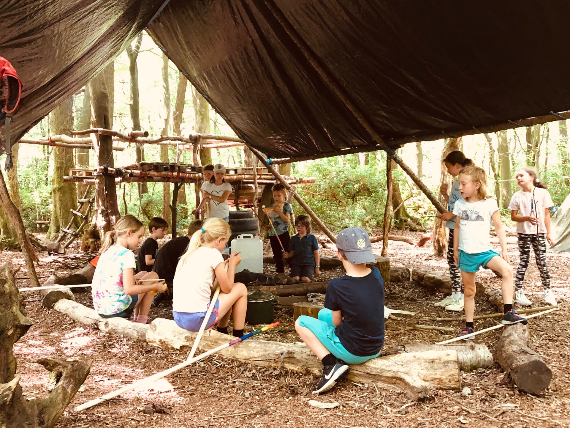Children and instructors under a tarpaulin shelter beside a fire circle in the Belmont School woodland base camp, with timber platform structures behind