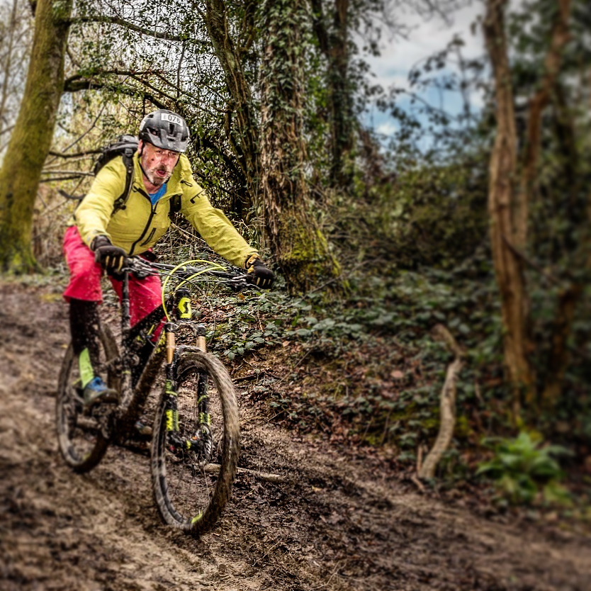 A rider in a yellow jacket riding a mountain bike down a wet, muddy woodland trail