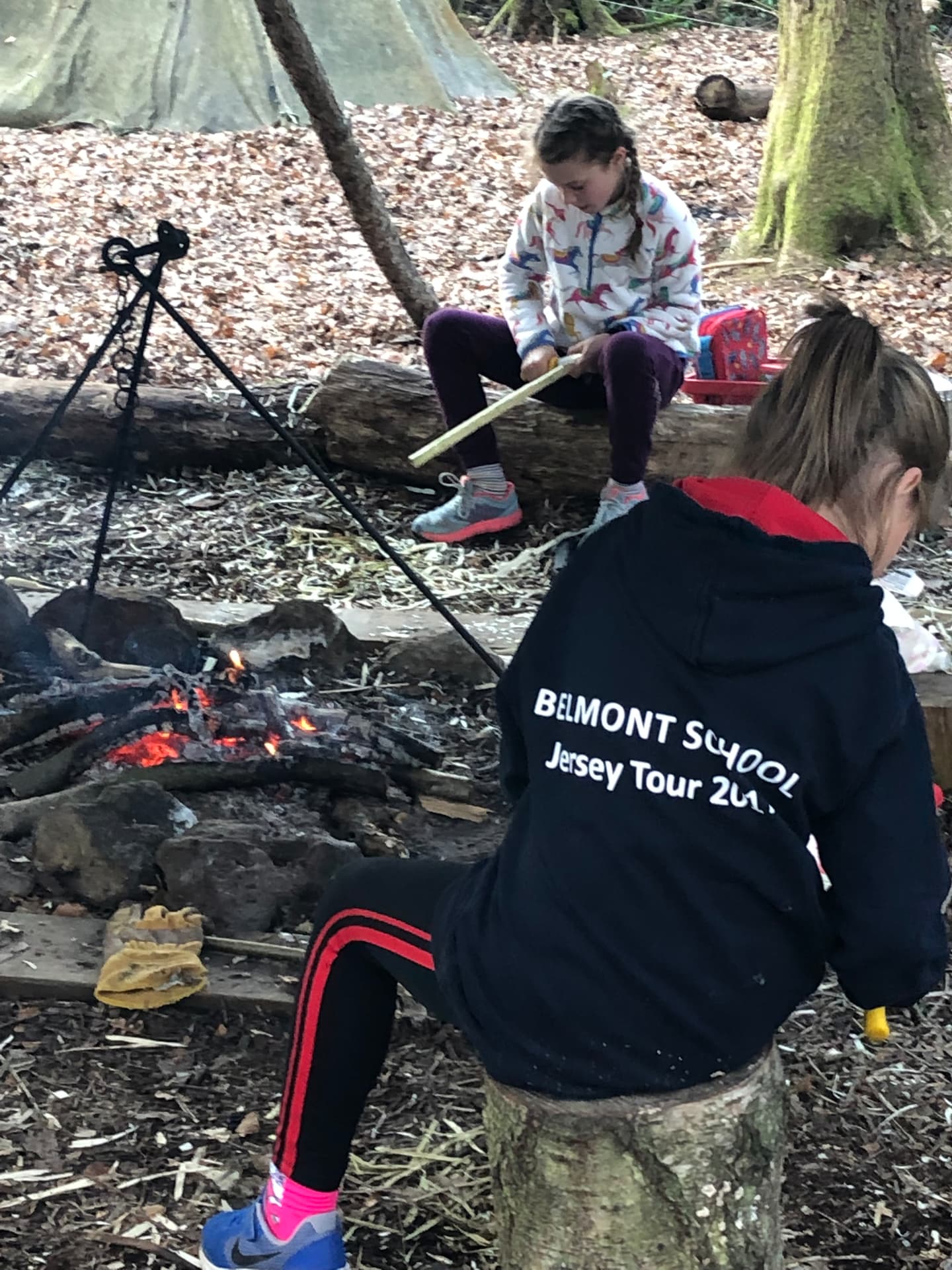 Two young people in Belmont School clothing carving wood beside a fire pit in the school woodland