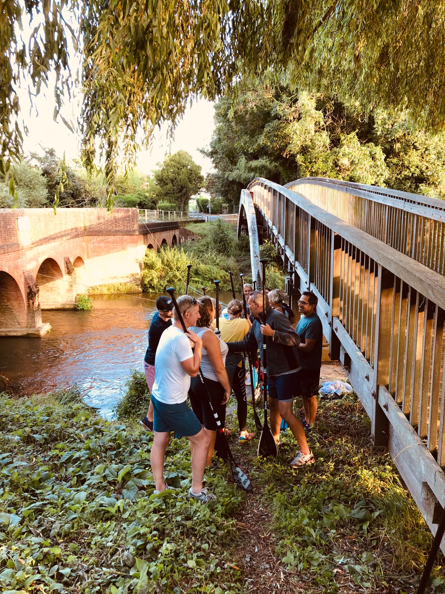 An organised group gathered with paddleboards and paddles beside the River Mole at a bridge, receiving a briefing before setting off
