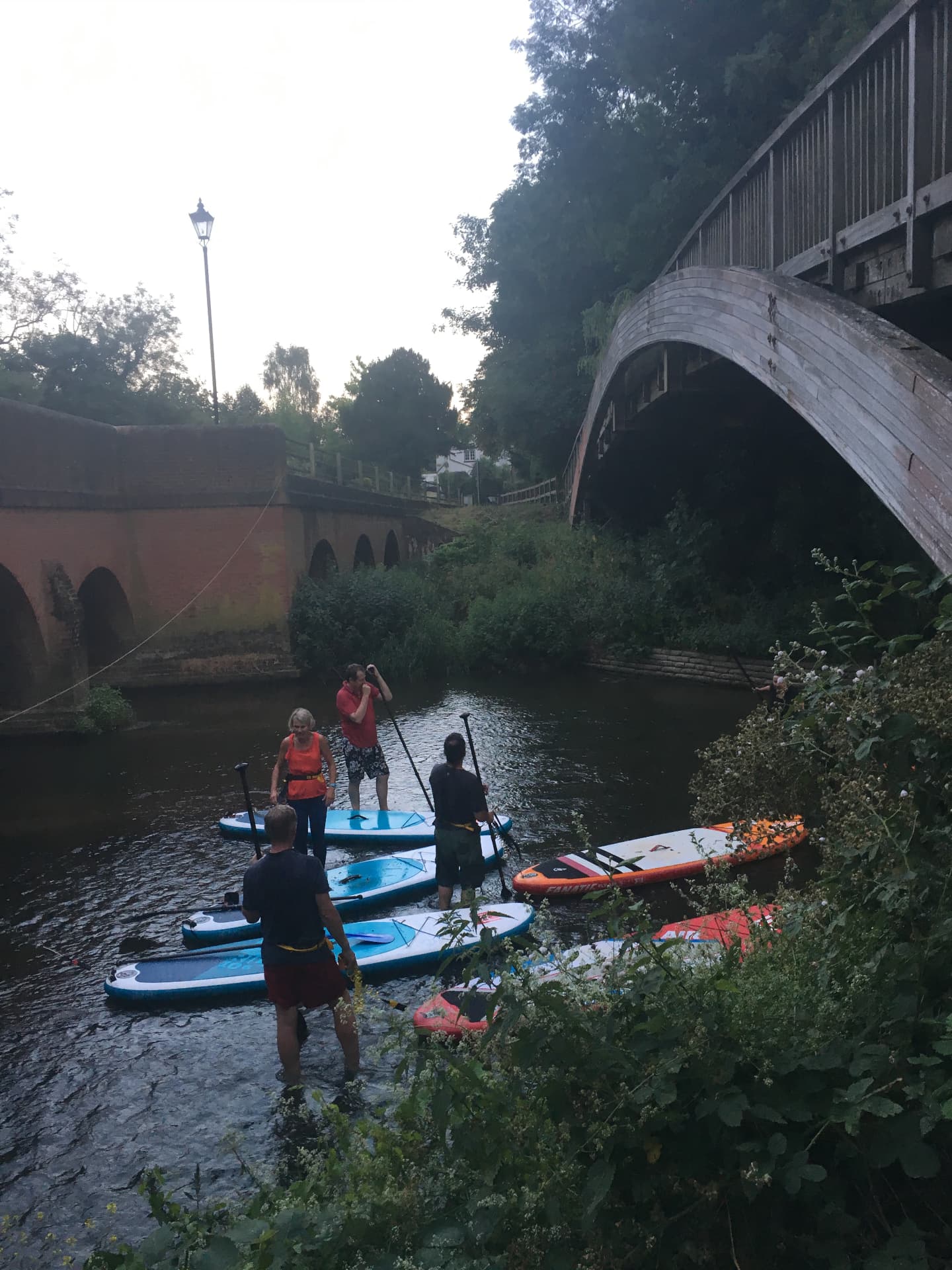 Paddlers launching and setting off on the River Mole beneath a timber arch bridge, with instructors standing in shallow water alongside the boards