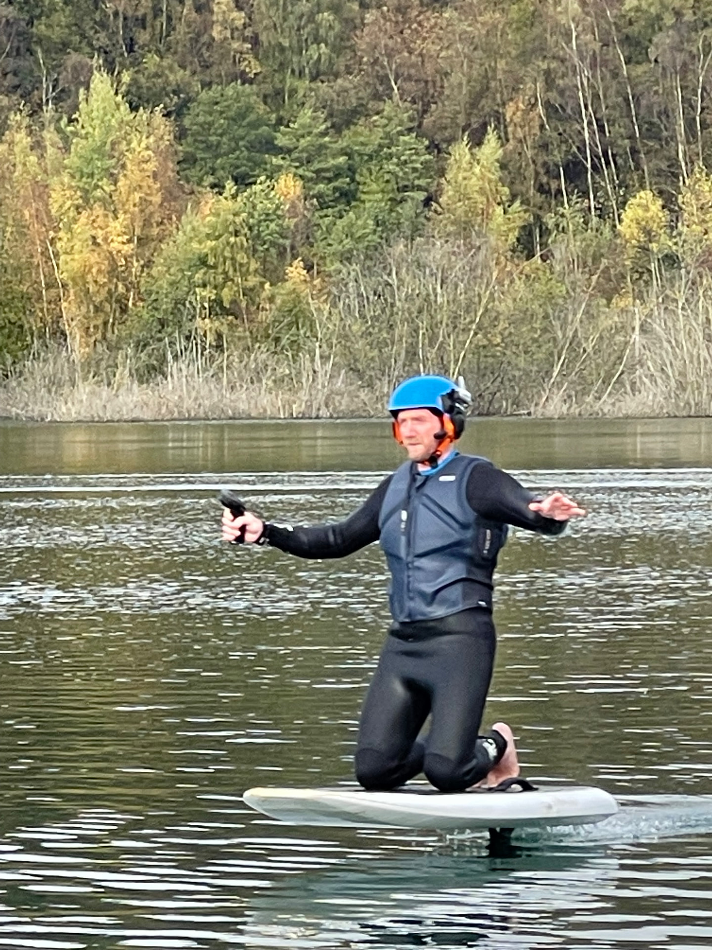 An e-foil rider progressing from kneeling control towards standing flight during instruction at Buckland Park Lake, arms out for balance with the board just lifting