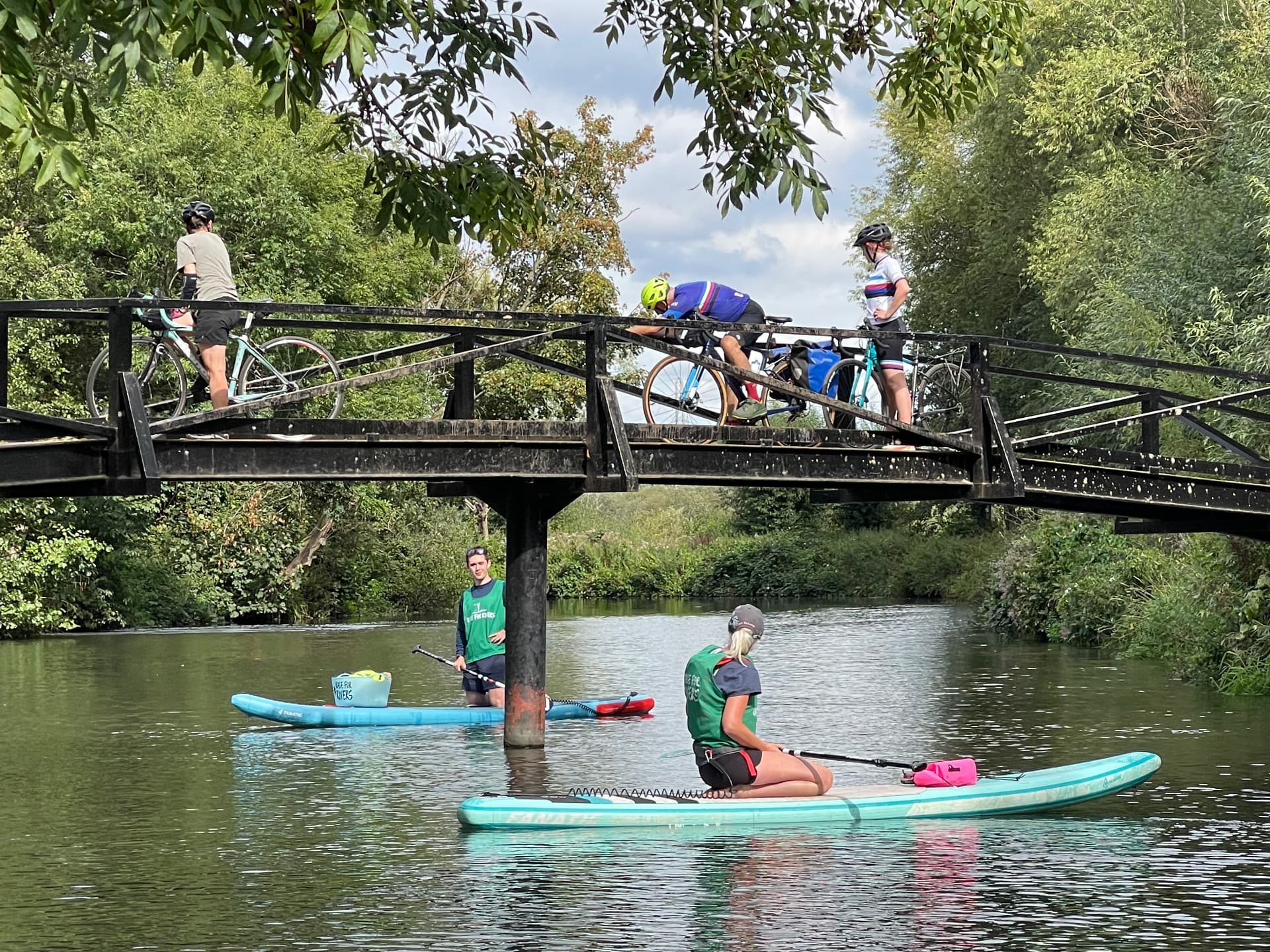 Two paddleboarders pause under a footbridge on the River Wey while cyclists cross above them on the towpath