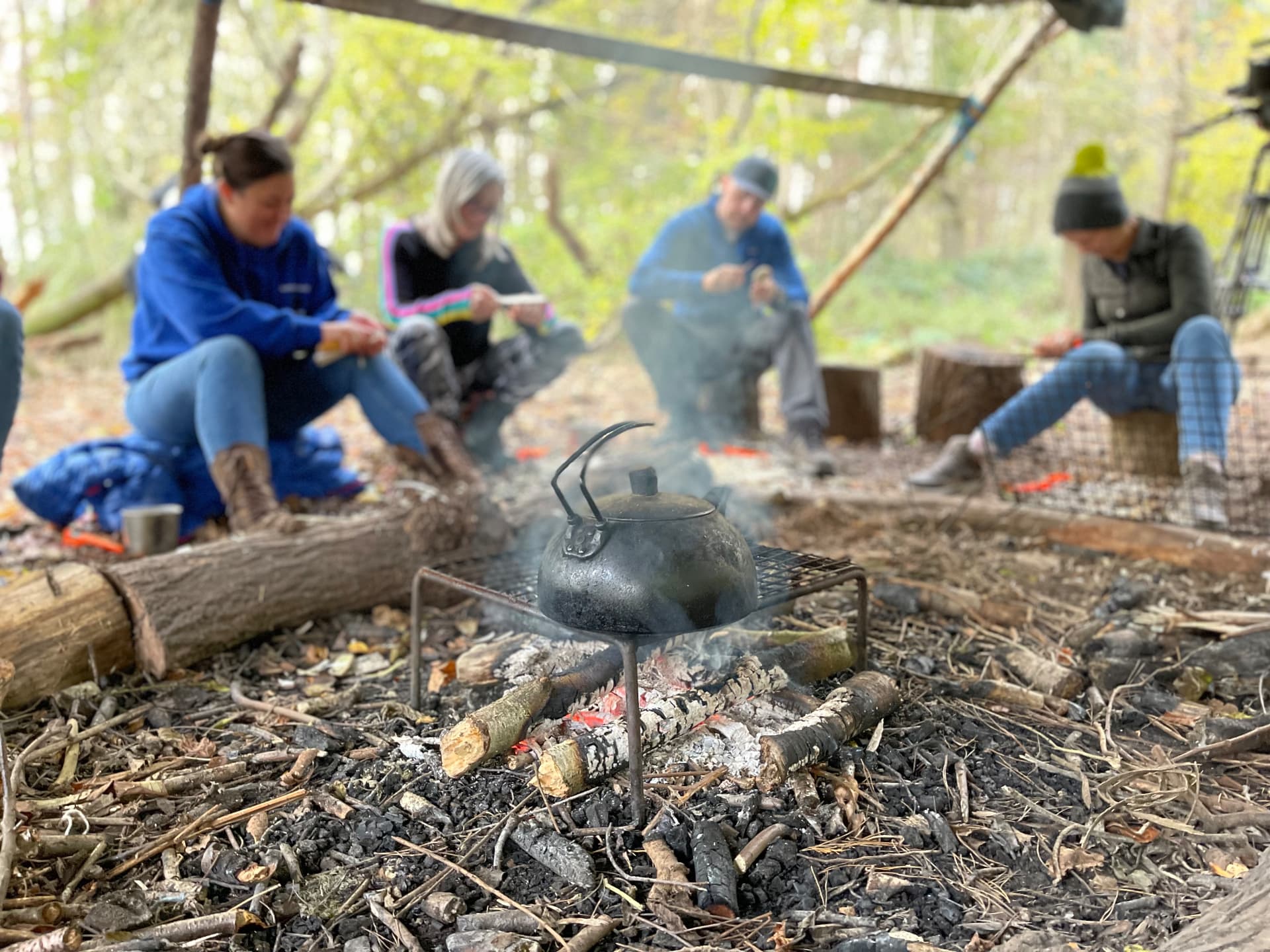 A woodland learning session around a small fire — kettle on a grate, group seated on logs working with hand tools under a tarp shelter