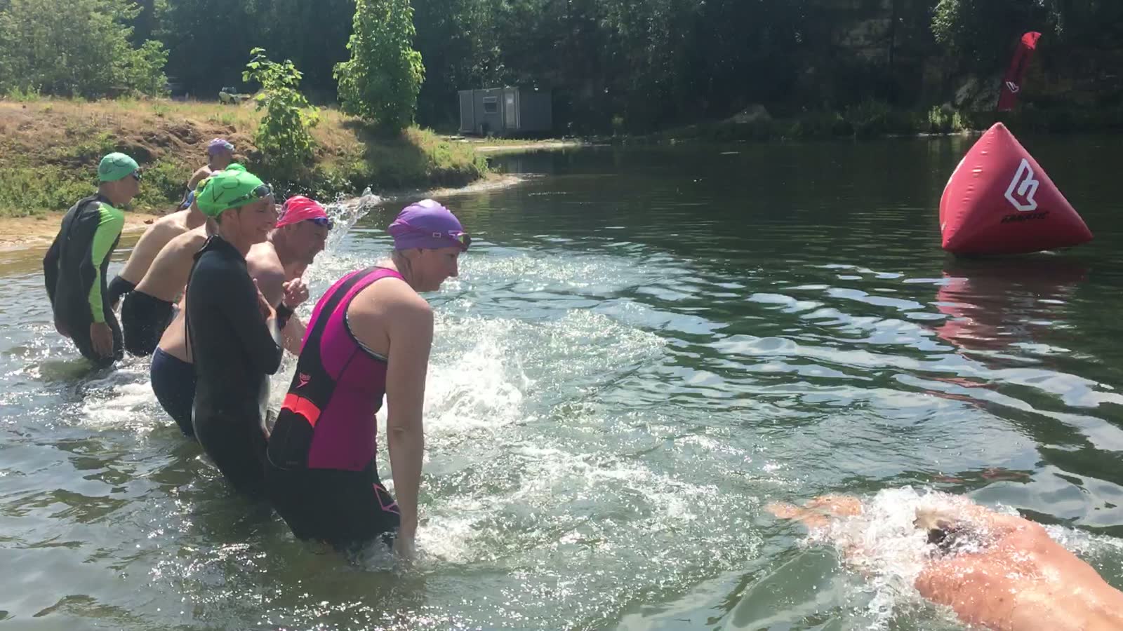 A coached group of open water swimmers in wetsuits entering an inland lake beside a marked course buoy in 2018