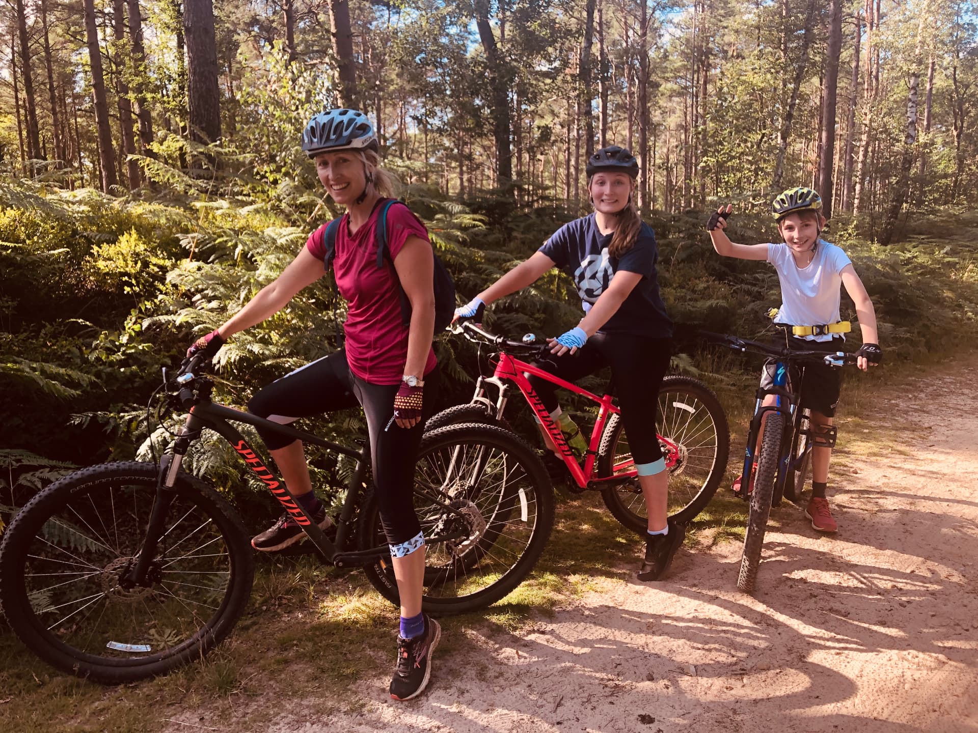 Three riders of different ages standing with mountain bikes on a sunlit sandy trail surrounded by ferns and pine trees