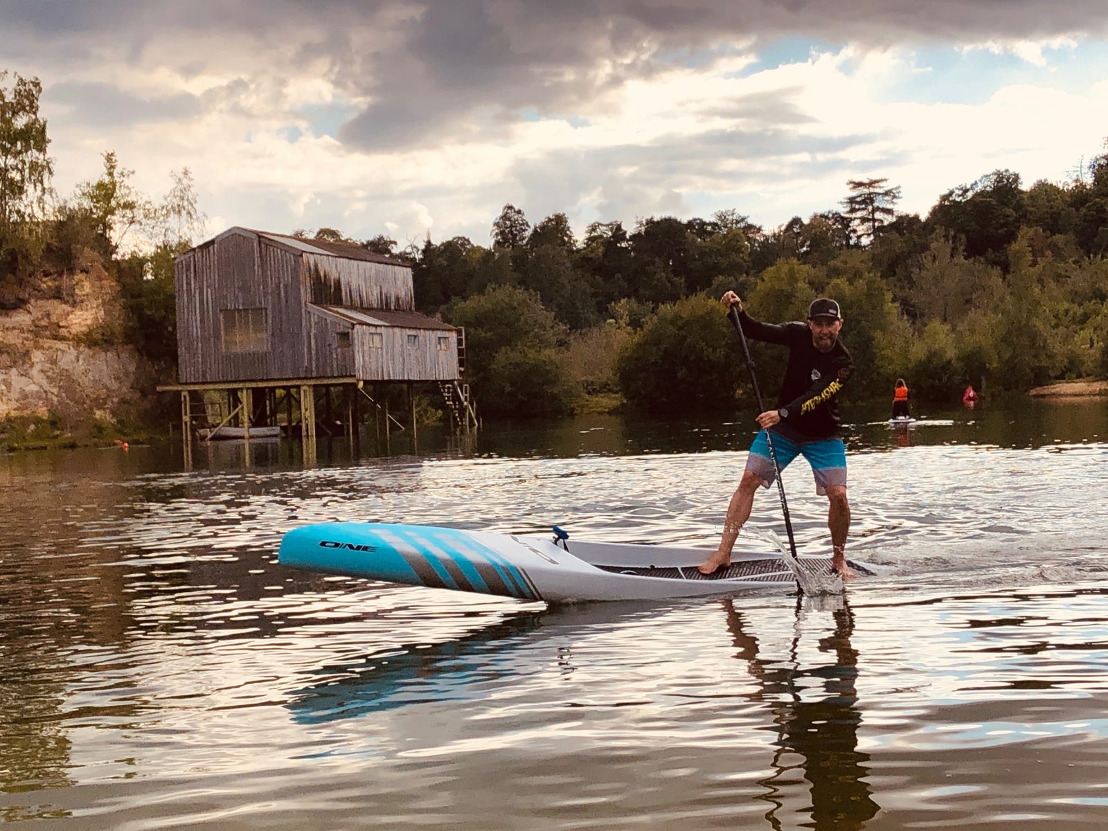 Paddler driving a long touring board forward on still water in the evening, past an old timber boathouse