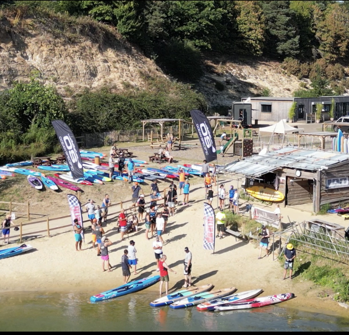 A busy outdoor activity venue in operation — boards laid out on the beach, event flags, equipment store and participants gathering by the water