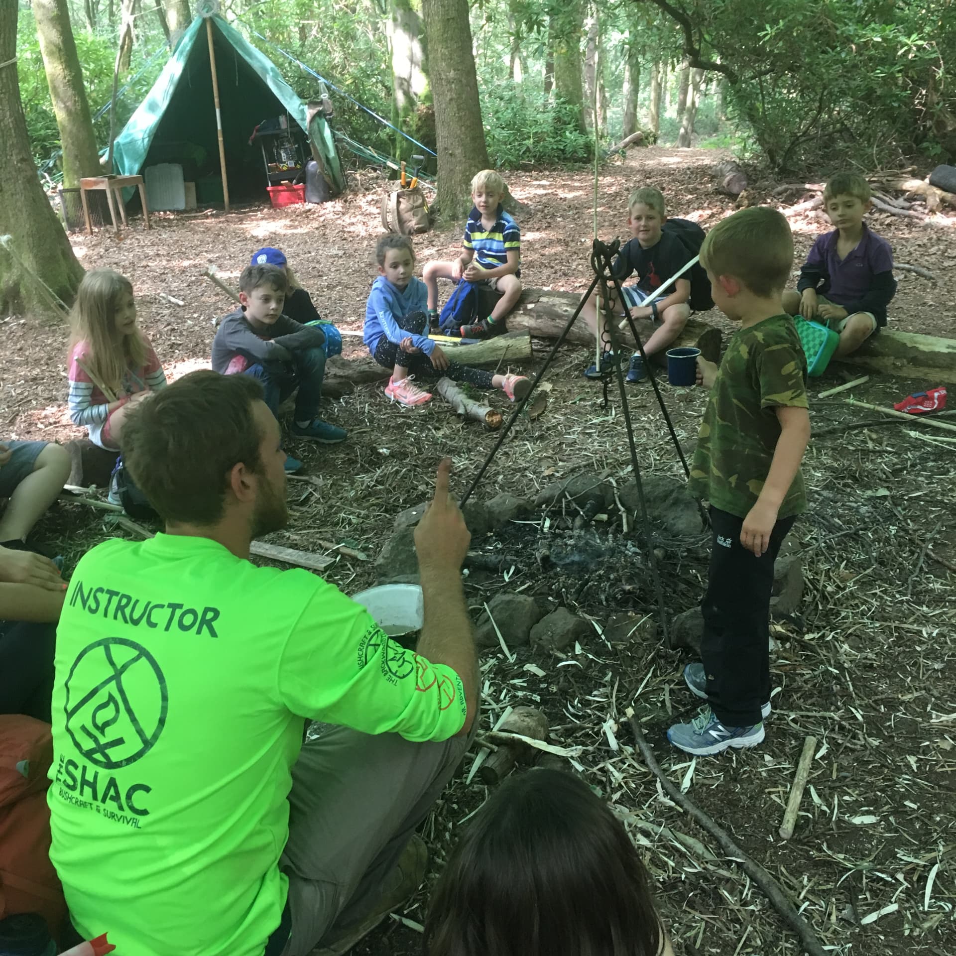 A Surrey Hills Adventure Company instructor in a high-visibility top talking with a seated group of children around a woodland fire pit