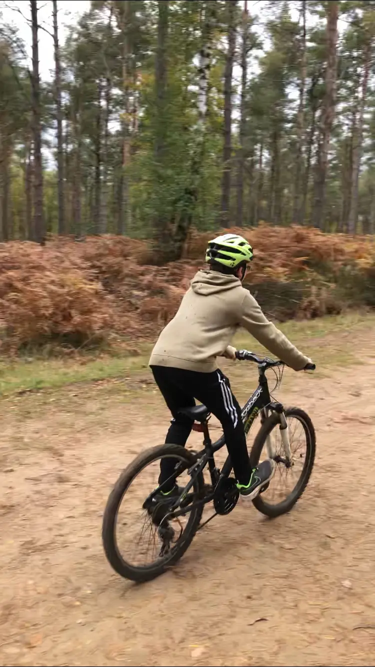 A young rider in a helmet riding a mountain bike along a wide sandy fire road through pine woodland