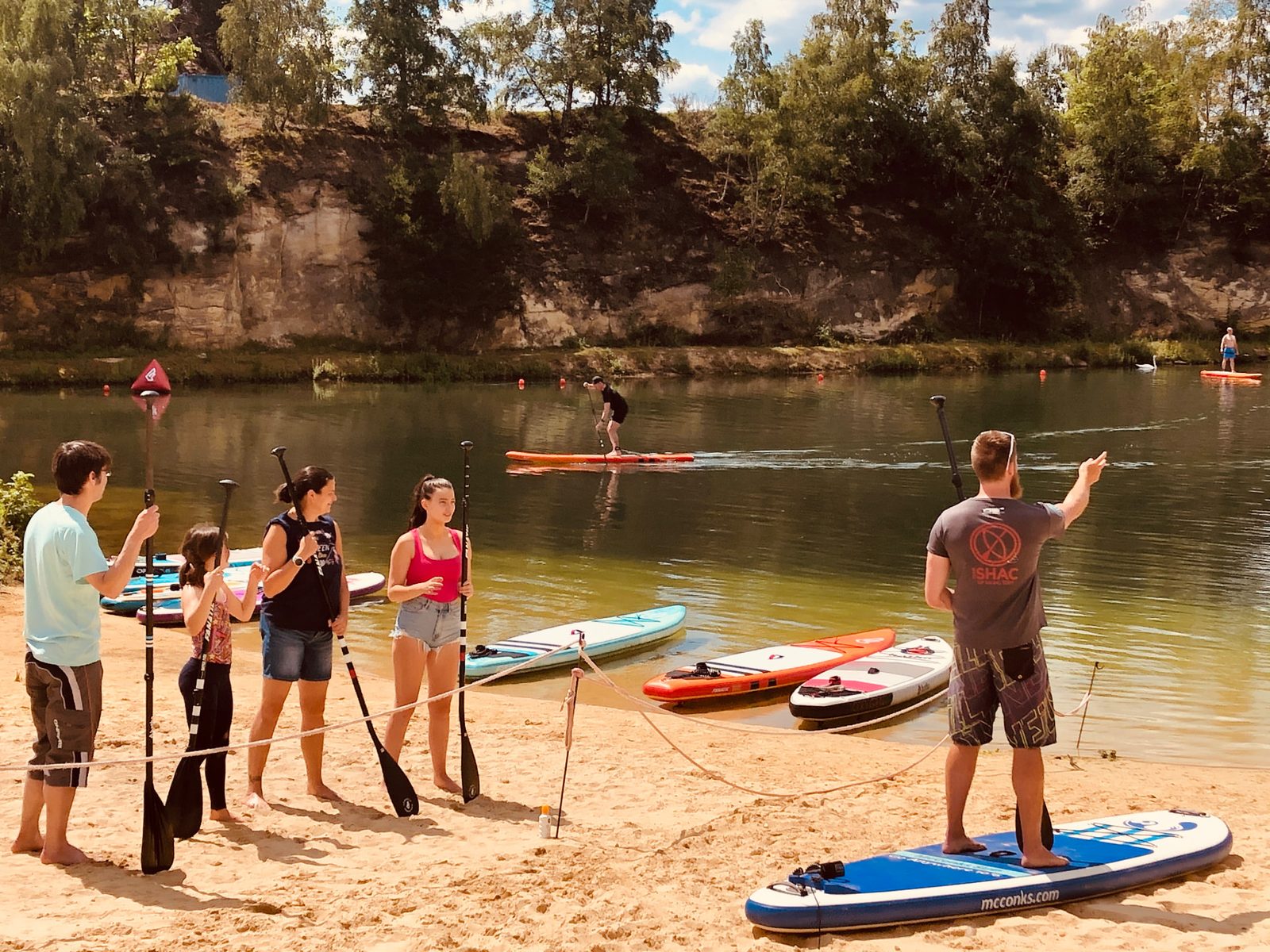Instructor briefing a small group of paddleboarders holding paddles on a sandy lake shore beside their boards