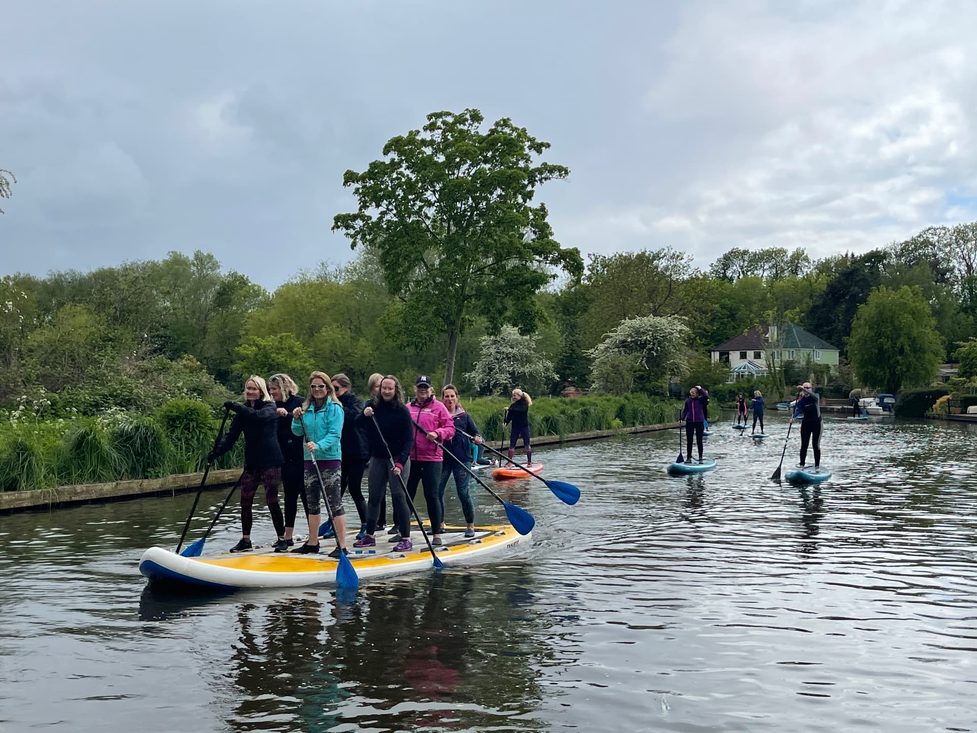 A large group of paddlers standing together on an oversized multi-person paddleboard on the River Wey, with other paddlers following behind