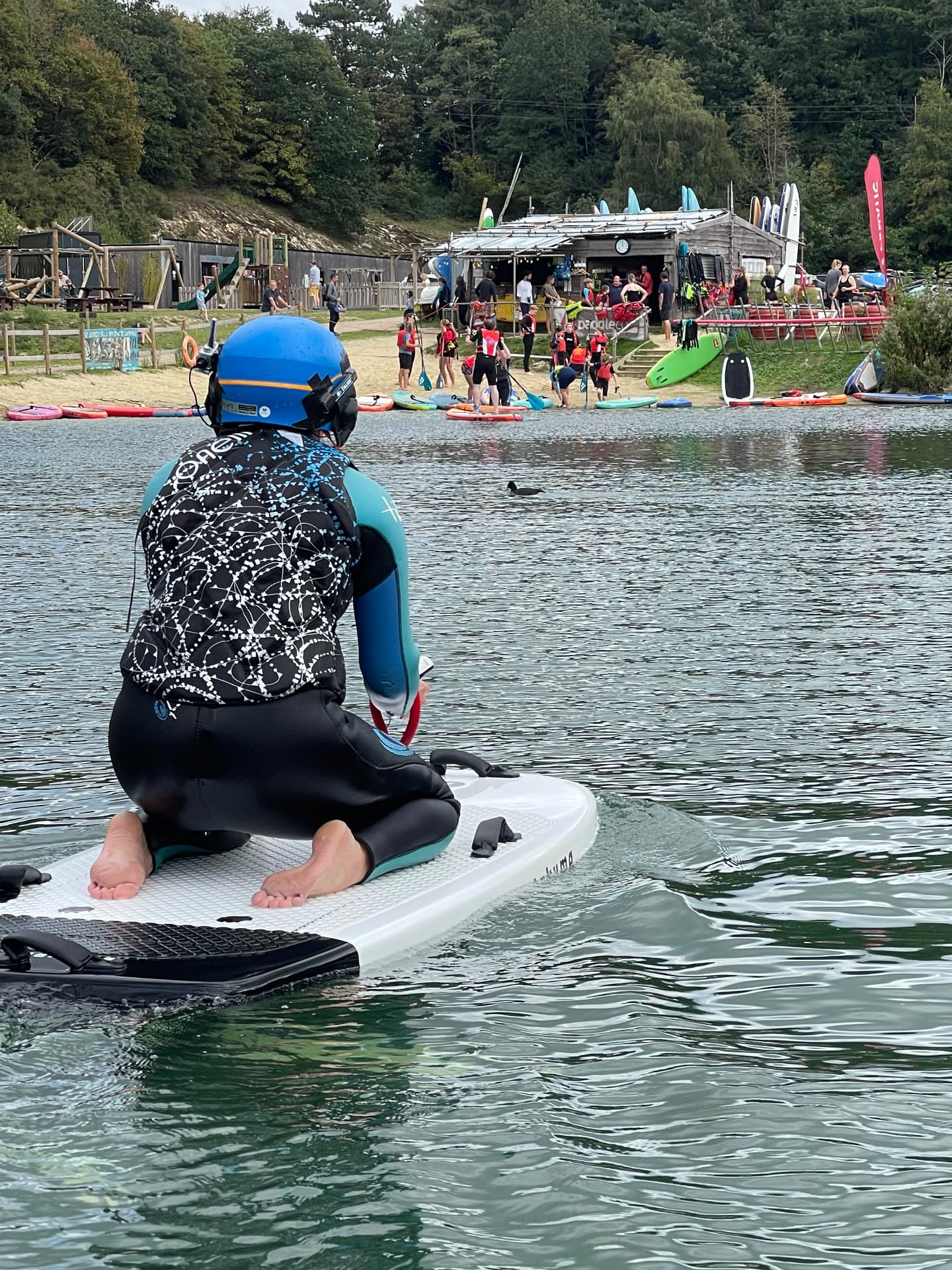 An e-foil rider kneeling low on the board, controlling the throttle while looking towards the beach and activity centre at Buckland Park Lake