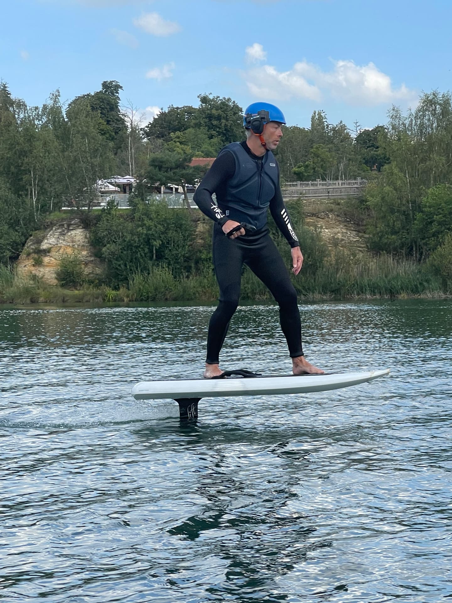 An e-foil rider standing on the board and flying clear of the water on a summer day at Buckland Park Lake, the foil mast fully visible beneath the board