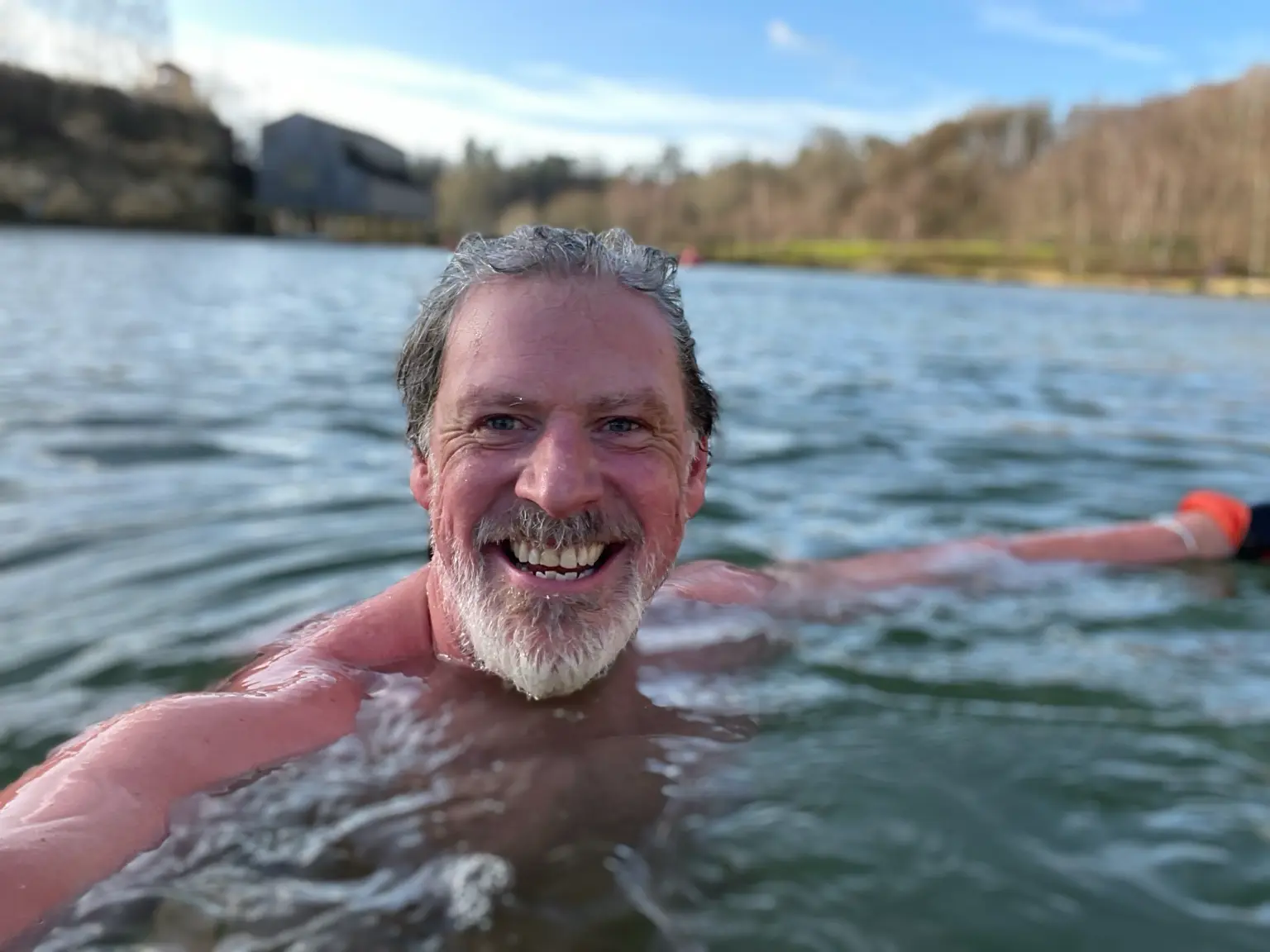Skip swimming in a cold inland lake, smiling at the camera with the shoreline behind him
