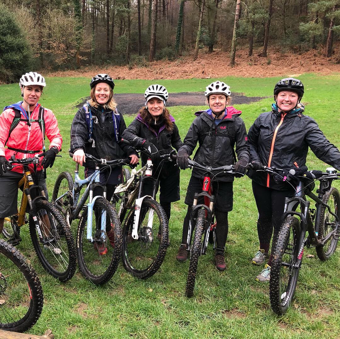 A group of five adult riders in waterproof jackets standing with muddy mountain bikes on wet grass in front of woodland