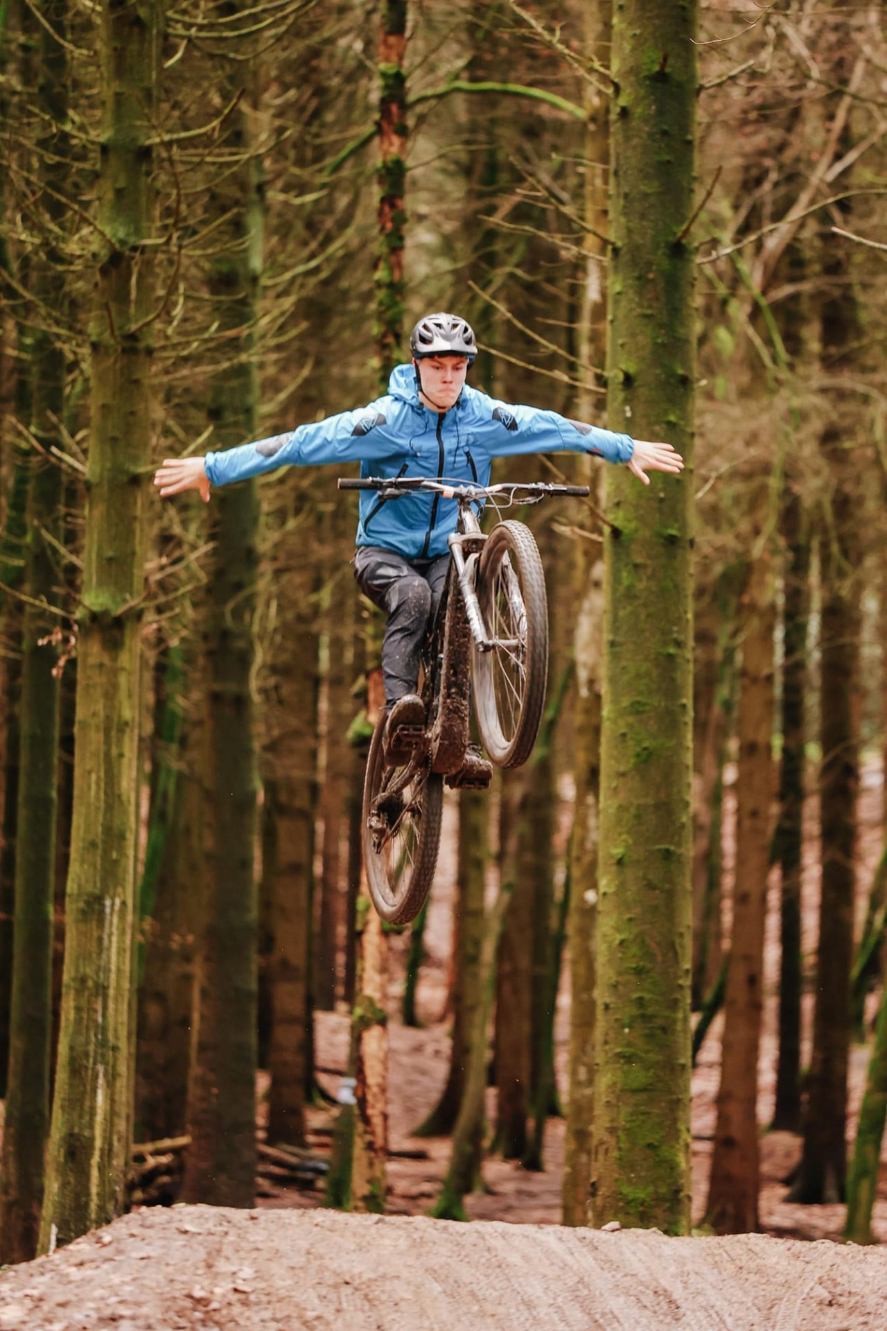 A mountain biker in a blue jacket airborne above a dirt jump in a mossy pine woodland, arms outstretched from the handlebars
