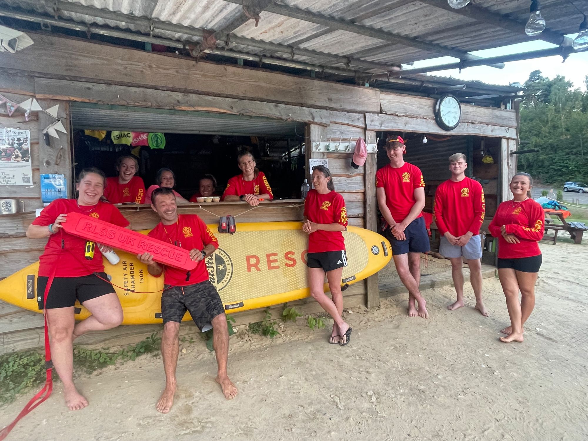Open-water lifeguard team in red kit with a rescue board and radios outside the lifeguard hut at an inland lake venue
