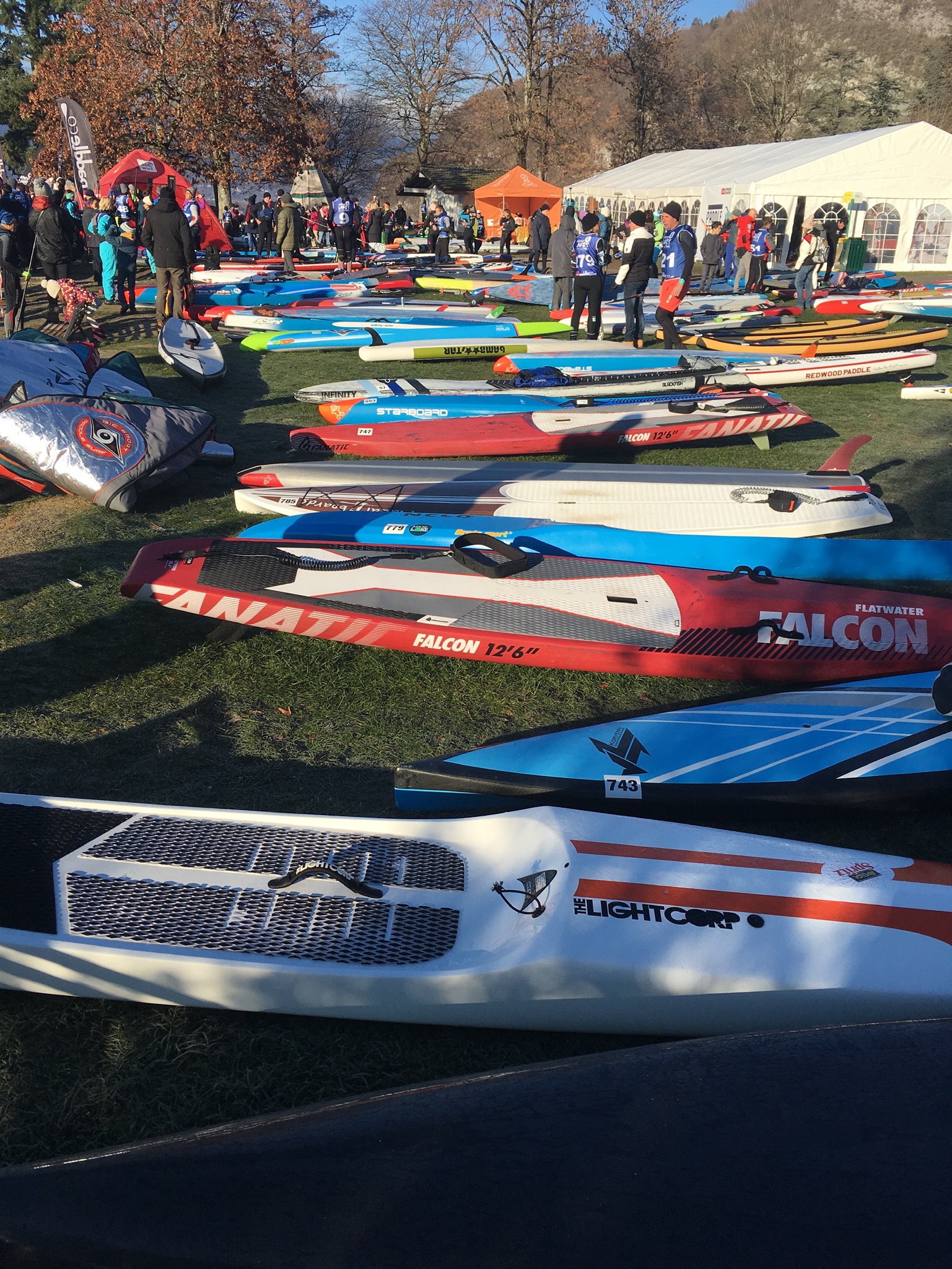 Rows of race paddleboards laid out on frosty grass with paddlers gathering by event tents before a winter SUP race in France