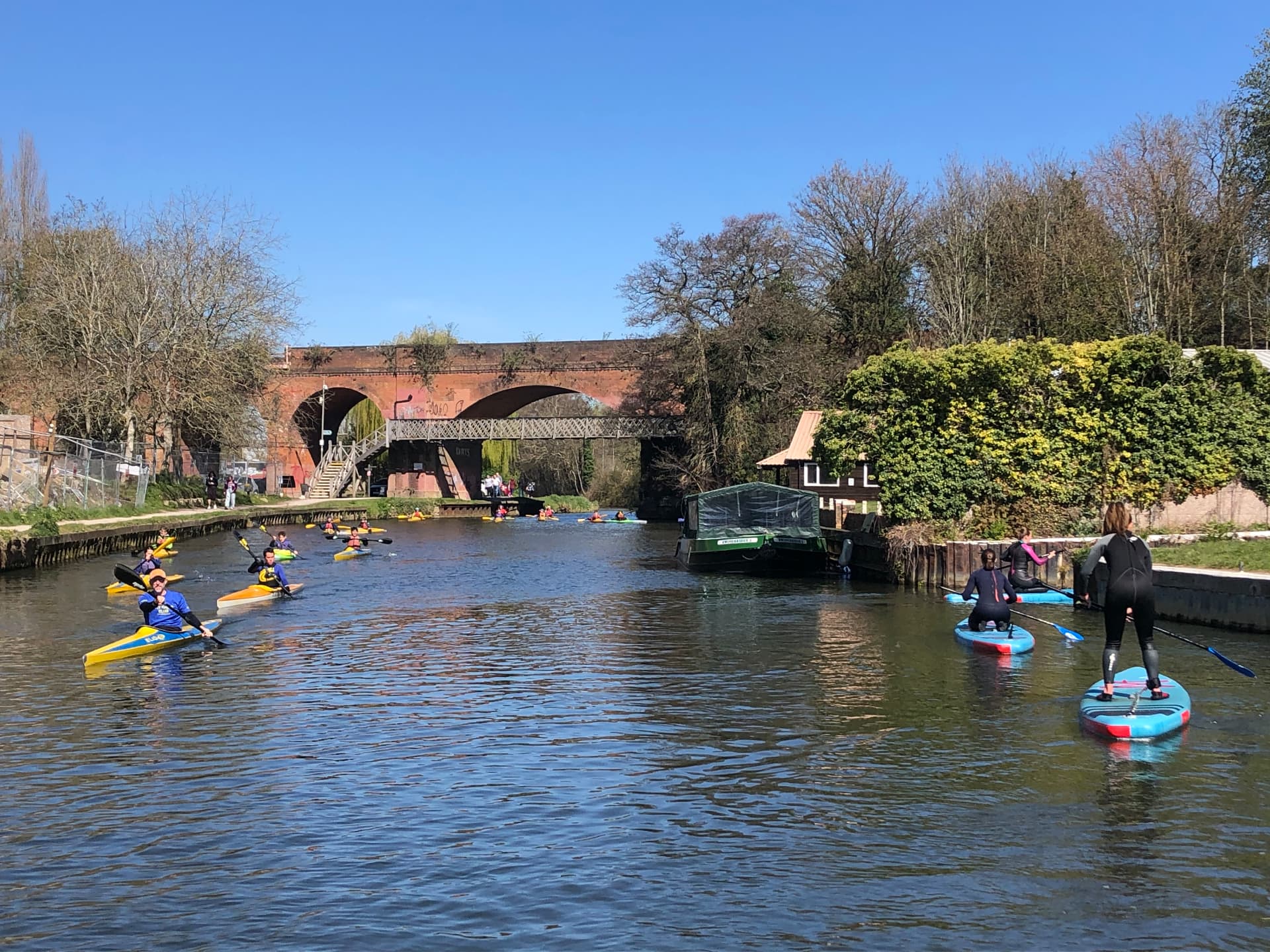 Paddleboarders and a group of kayakers sharing the River Wey Navigation near Guildford, passing a boathouse beneath a brick railway viaduct