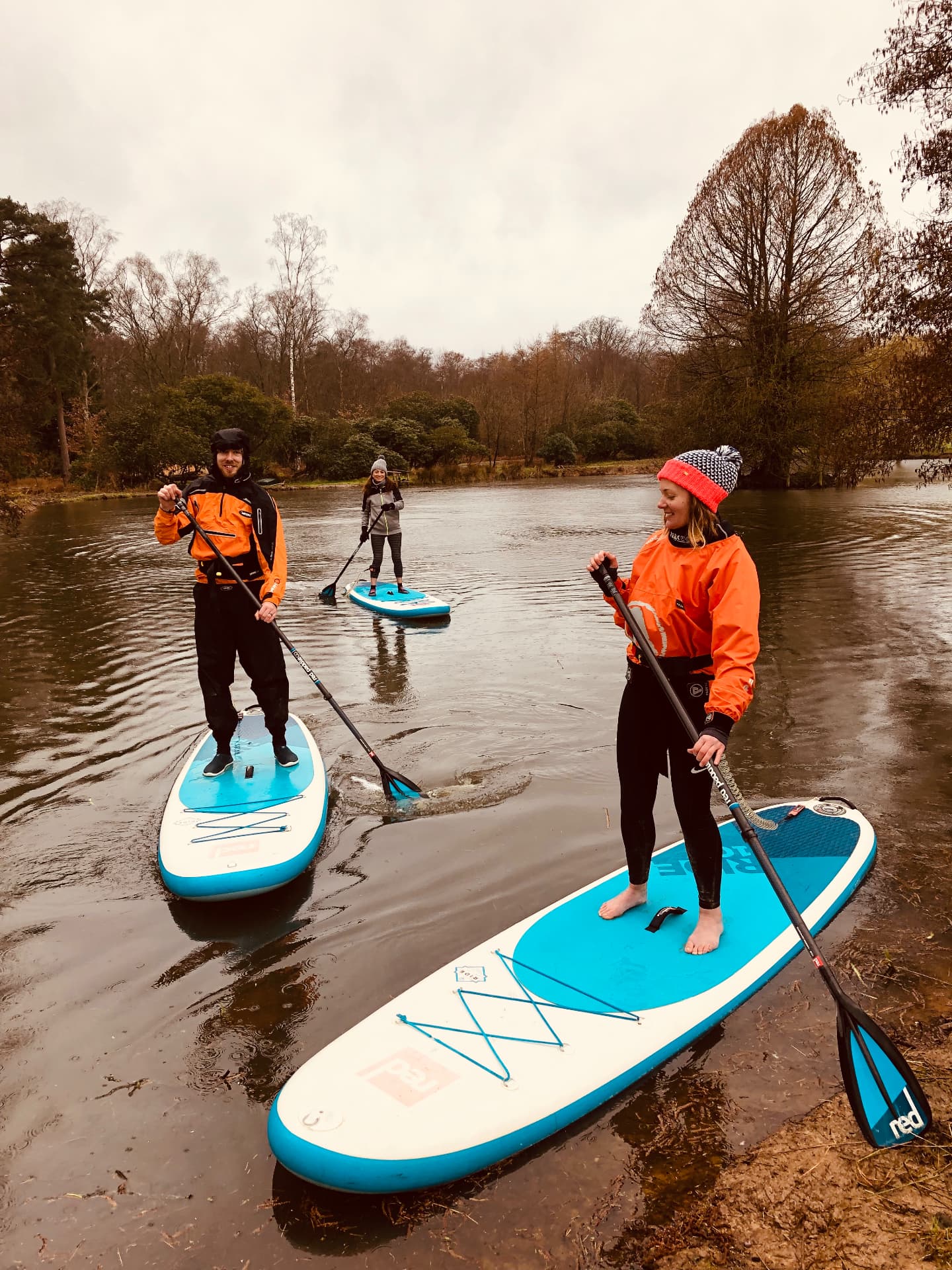 Three paddleboarders in drysuits and buoyancy aids on flat sheltered water surrounded by woodland