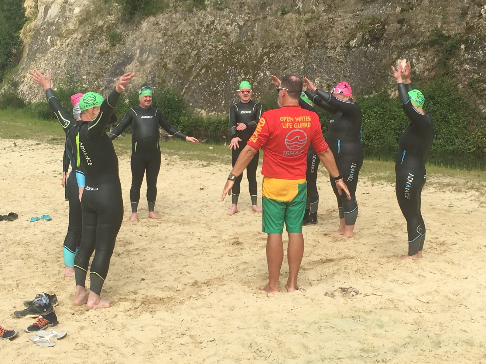 Scott Innes in an open water lifeguard top briefing a group of swimmers in wetsuits on the beach before a swim